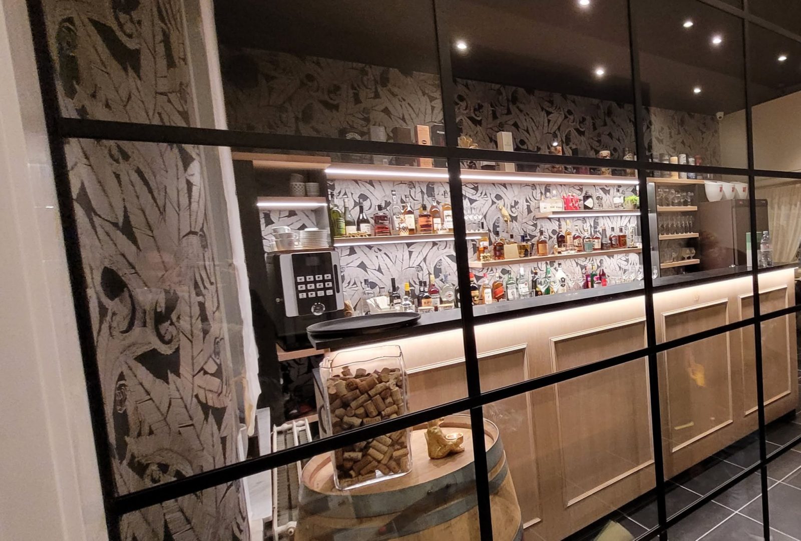 Le comptoir du bar et ses étagères de bouteilles rétroéclairées, derrière la verrière à cadres noirs