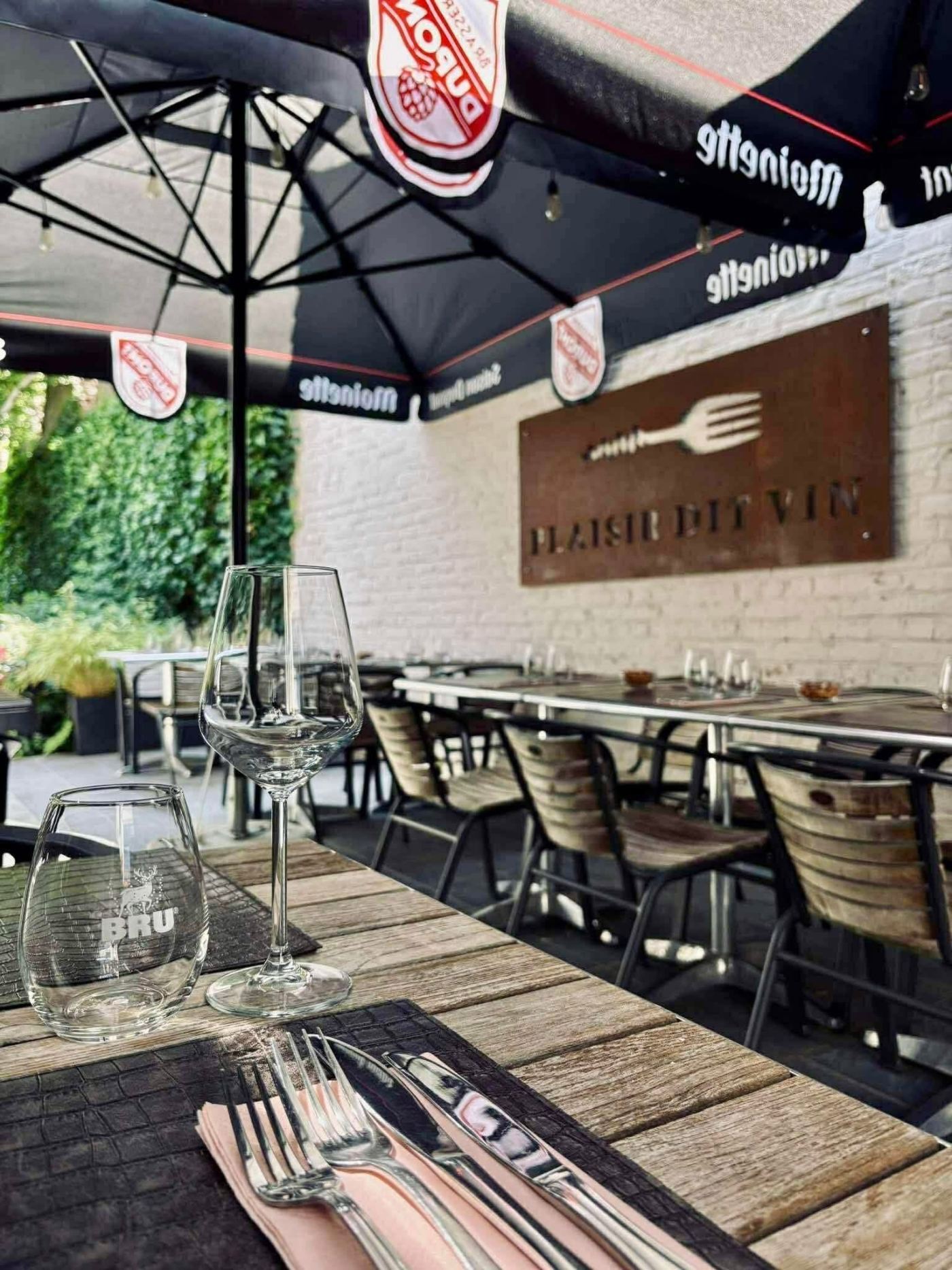 La terrasse du restaurant sous ses parasols, table en bois dressée et enseigne Plaisir dit Vin en métal rouillé sur le mur blanc